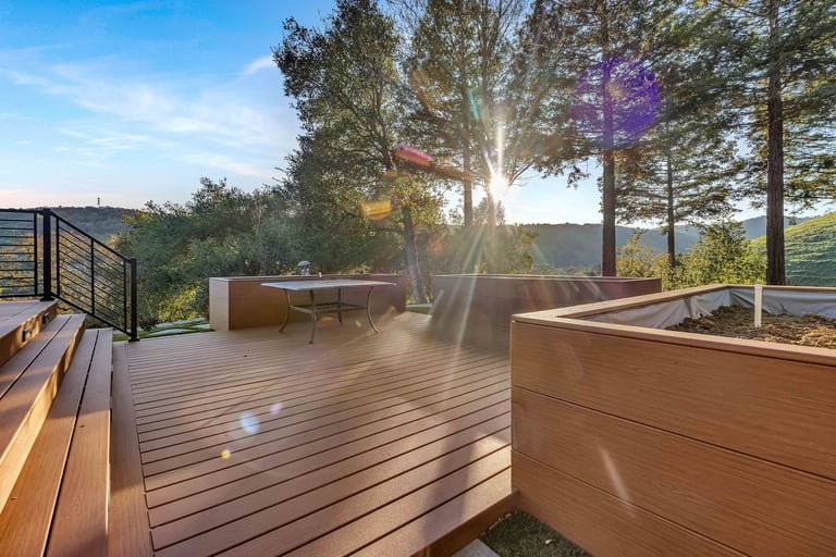 Modern wooden deck with sunlight streaming through tall trees, featuring wooden planters and metal railings overlooking a hillside view