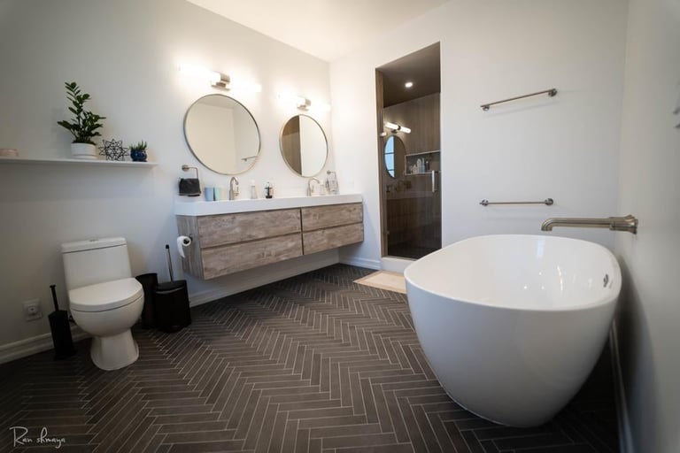 Modern bathroom with white freestanding tub, double vanity with mirrors, toilet, and herringbone tile flooring