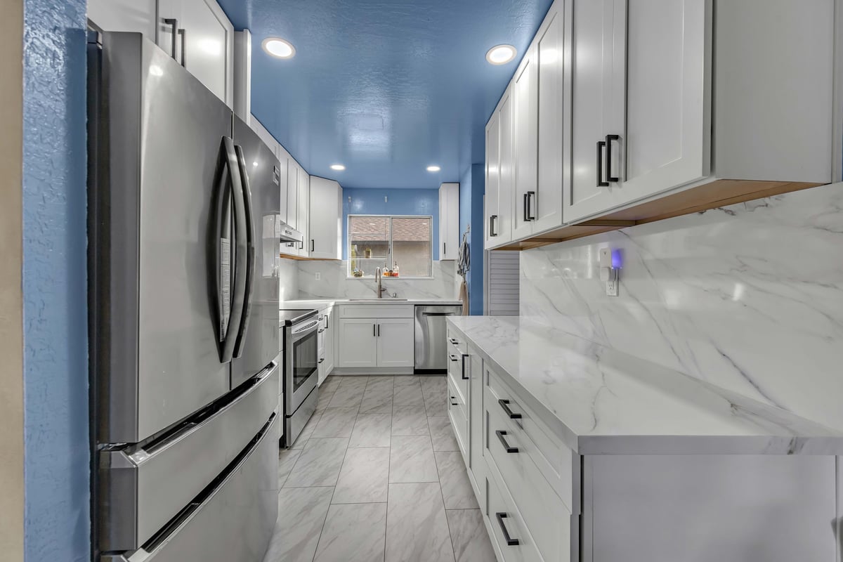 Modern galley kitchen with white cabinets, blue ceiling, marble countertop, and stainless steel appliances after renovation
