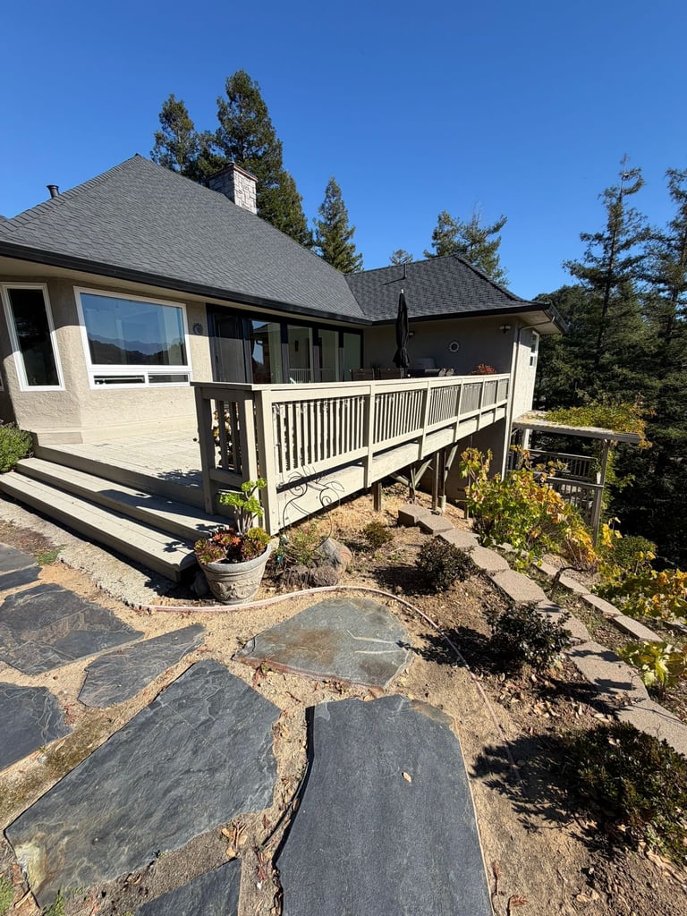 Modern home with cream and dark stone exterior, white railing deck, slate pathway, surrounded by evergreen trees under clear blue sky