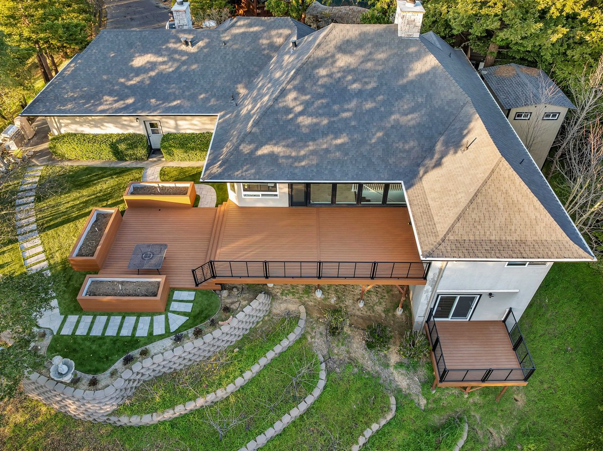 Aerial view of a modern home with copper roof, wooden deck, curved stone pathway, and manicured lawn surrounded by trees