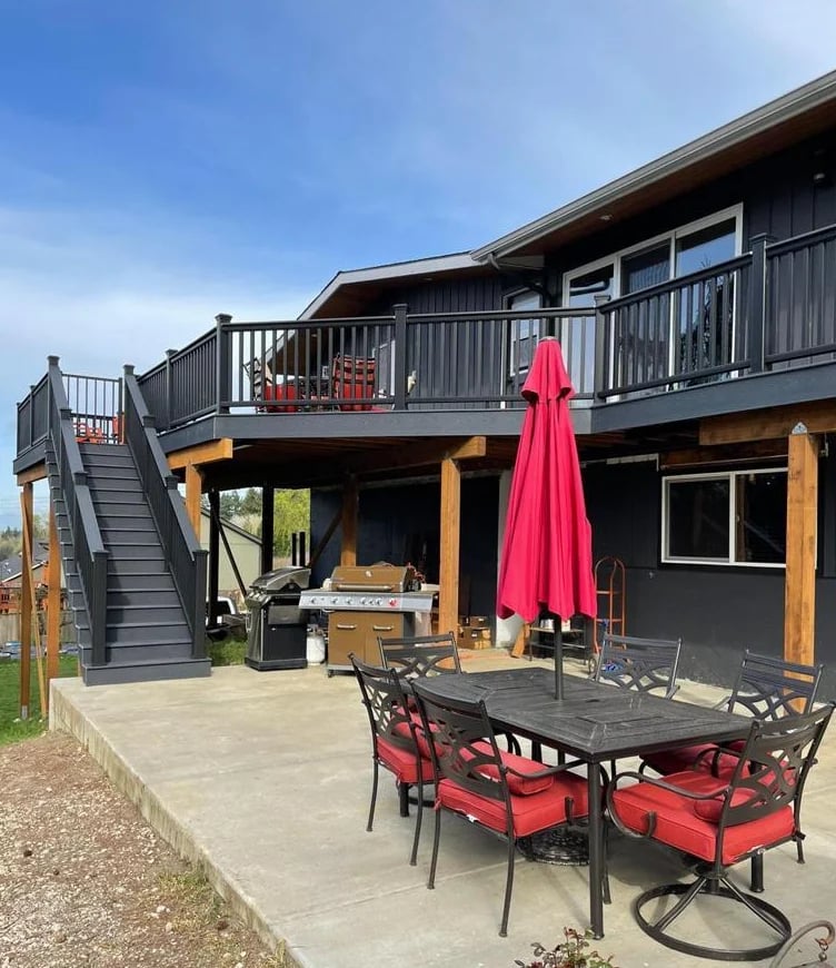 Modern two-story house with wooden accents, black railings, red patio furniture, and a red umbrella on a concrete patio