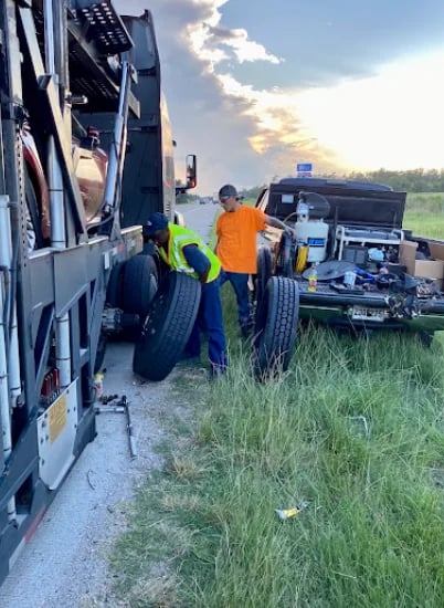 Roadside assistance workers in safety gear repairing a large truck tire on a highway with another vehicle nearby