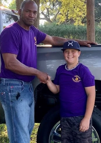 Adult man in purple shirt shaking hands with young boy in wheelchair wearing purple shirt and black cap