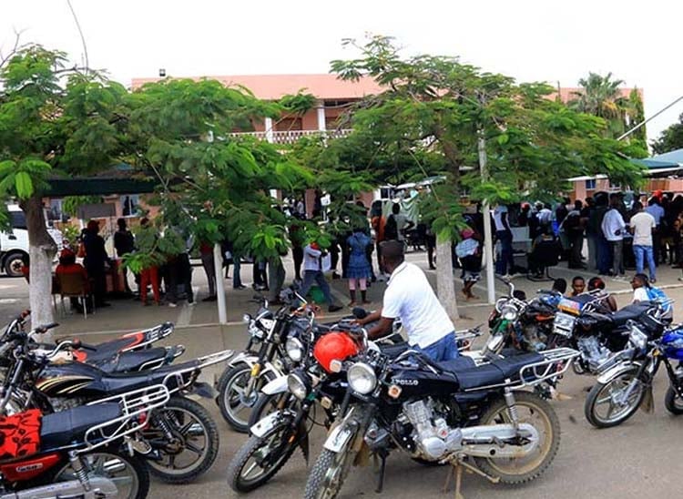 Motorcycles parked in foreground with crowds gathering under trees in an open-air market or community event setting