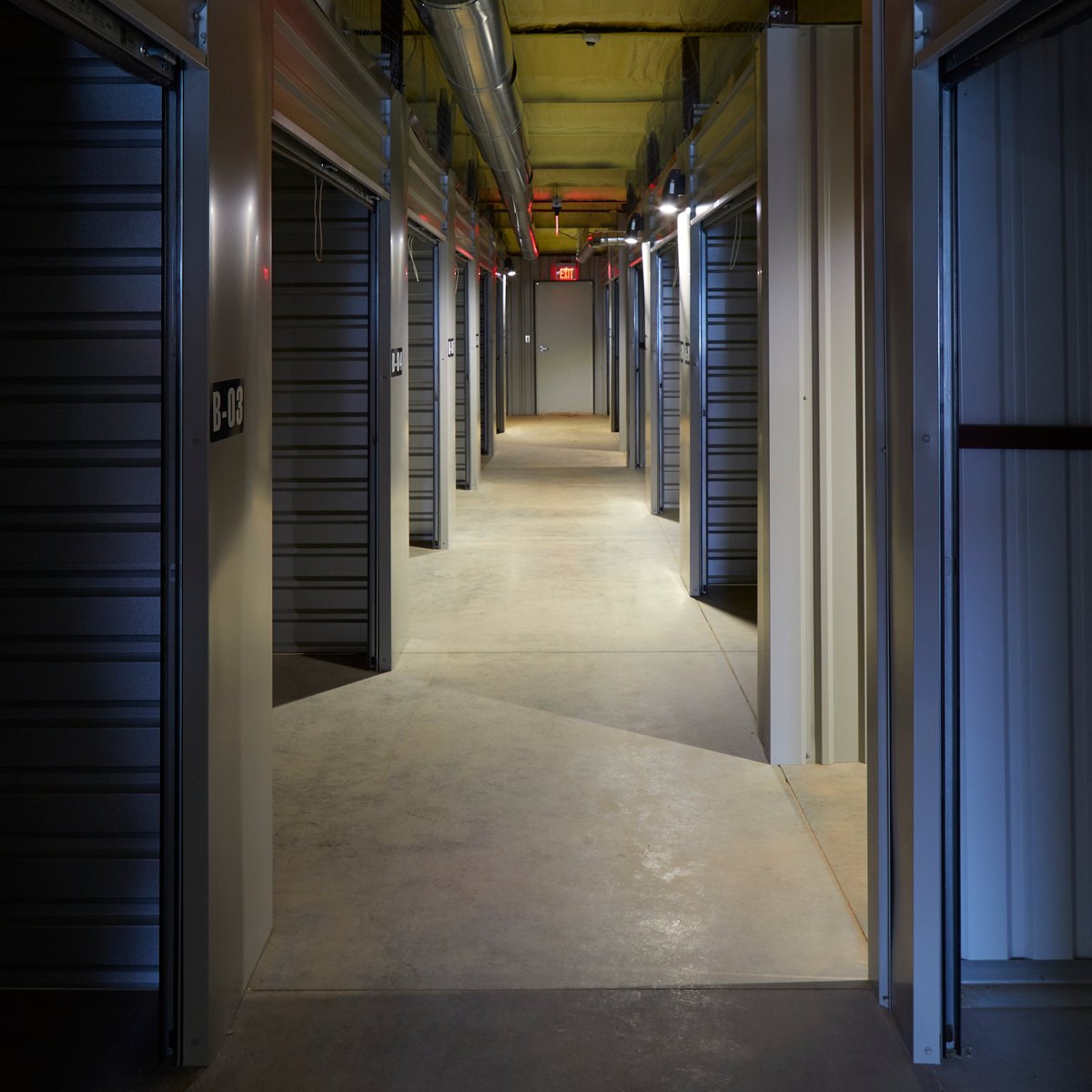 Long corridor of self-storage units with blue metal doors lining both sides, fluorescent lighting, and concrete floor