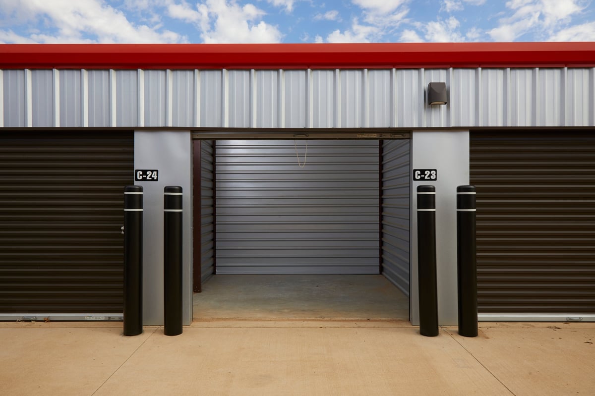 Front view of a Locked Up Self Storage unit with red roof, white and dark metal siding, open central garage door, and numbered units on either side