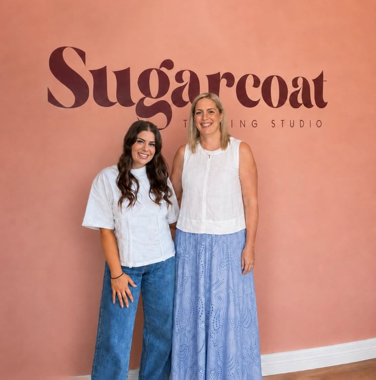 Two women smiling at Sugarcoat Waxing Studio, standing against a coral pink wall with the studios logo visible behind them