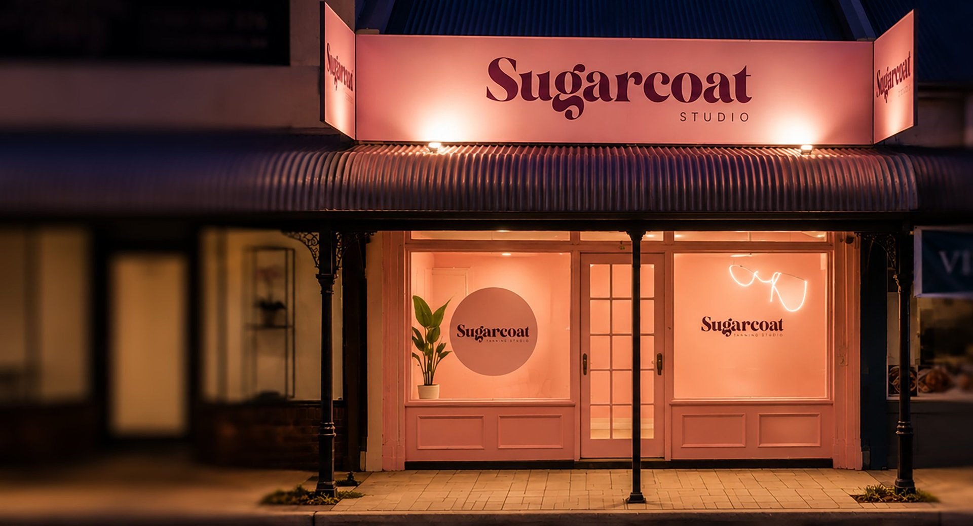 Sugarcoat Studio storefront at night with illuminated coral-pink awning and matching glass doors, minimalist storefront design