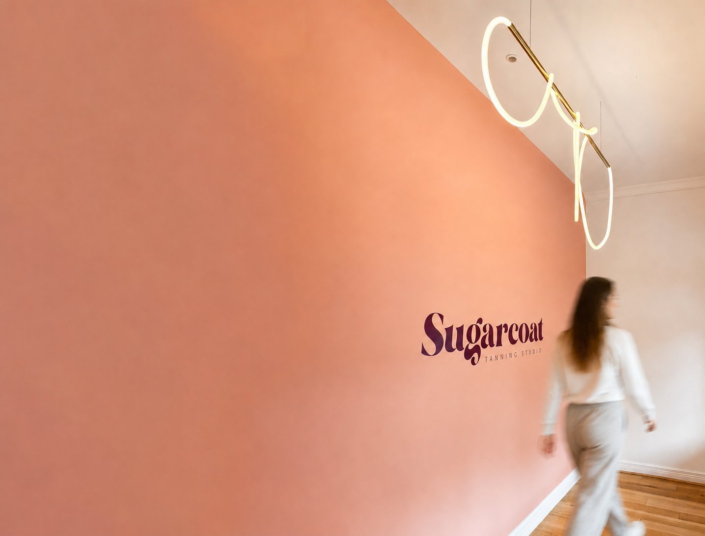 Sugarcoat salon interior with pink wall, illuminated glasses decor, and woman in white outfit walking past branding.