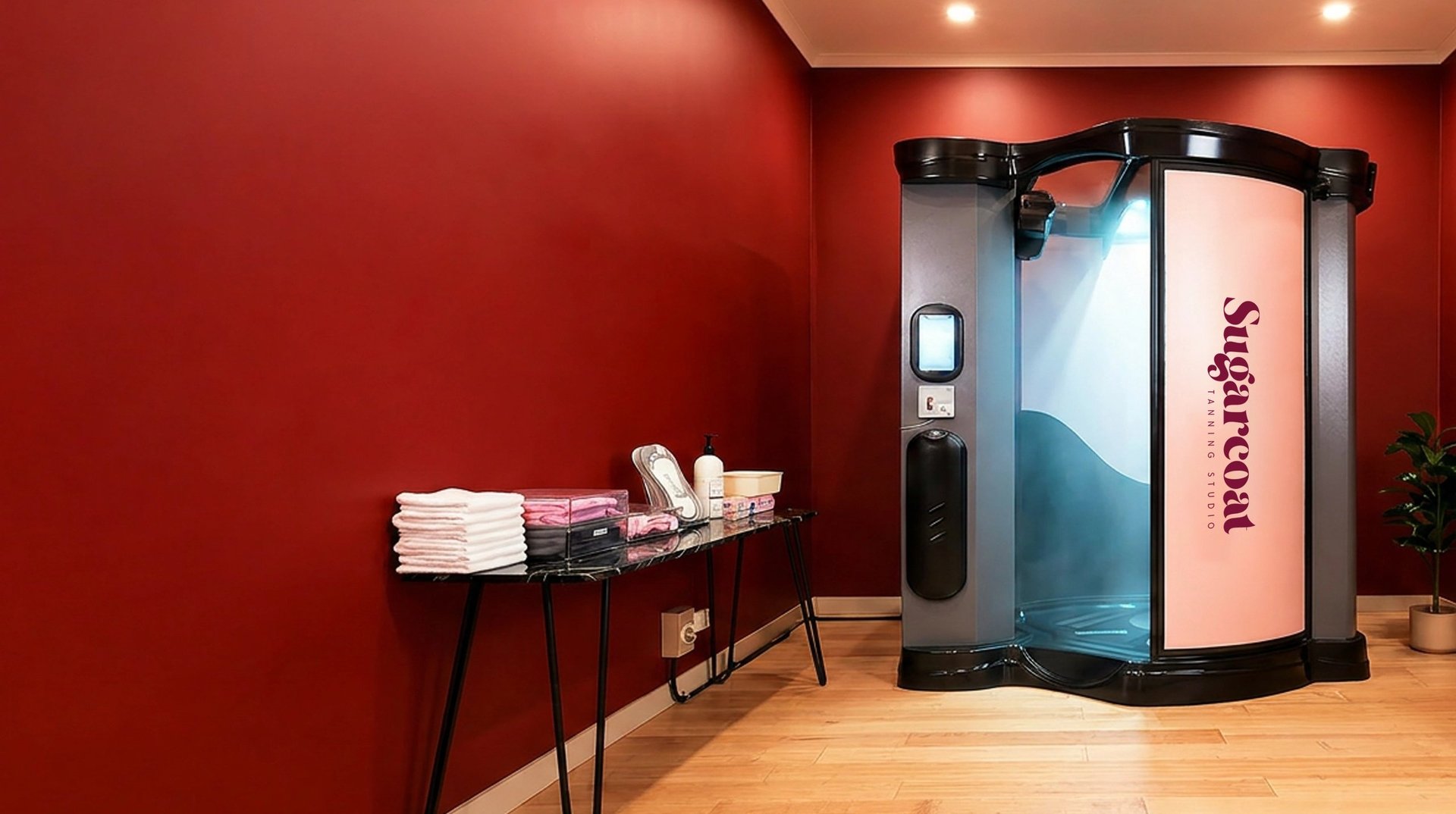 Modern spa interior with red walls, wooden floors, a Sugarcoat sauna booth with blue gradient panels, and a side table displaying white towels and toiletries