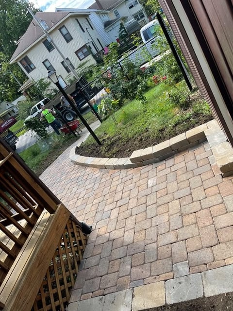 View from a deck overlooking a brick patio with raised garden beds and landscaping, with a residential house and yard visible
