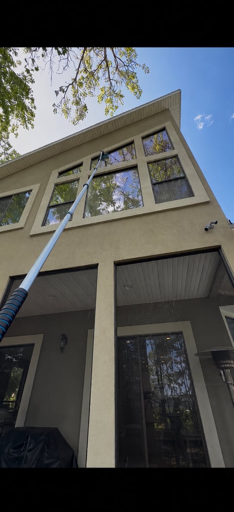 A two-story modern home with beige siding and large windows, being cleaned with a long-handled pressure washing pole