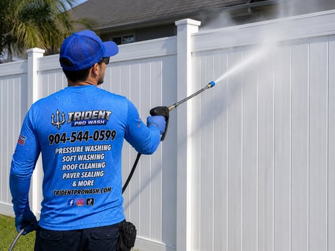 Worker in blue uniform and cap pressure washing a white fence with extended pole sprayer