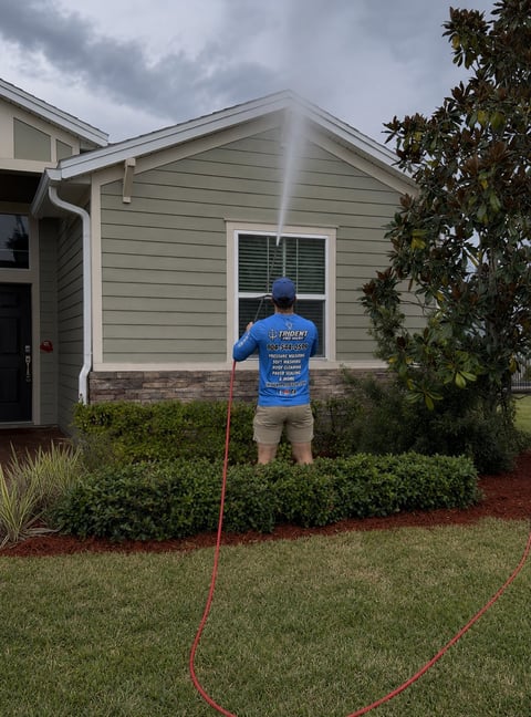 Person in blue jacket pressure washing the exterior of a residential home with a red hose