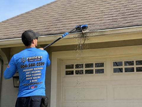Worker in blue shirt using pressure washer to clean roof gutters and eaves of residential house