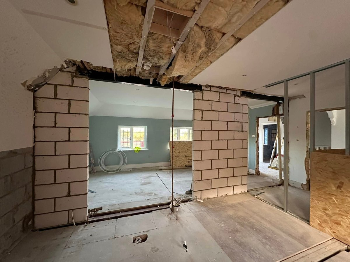 Interior construction site showing exposed brick walls, glass partition, wooden ceiling joists, and unfinished flooring