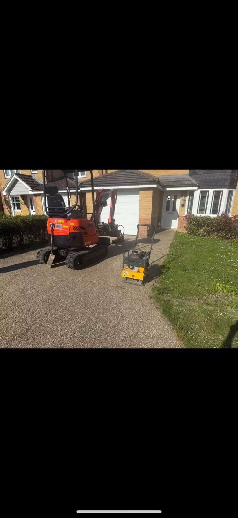 A red mini excavator breaking ground on an existing gravel driveway in front of a brick house