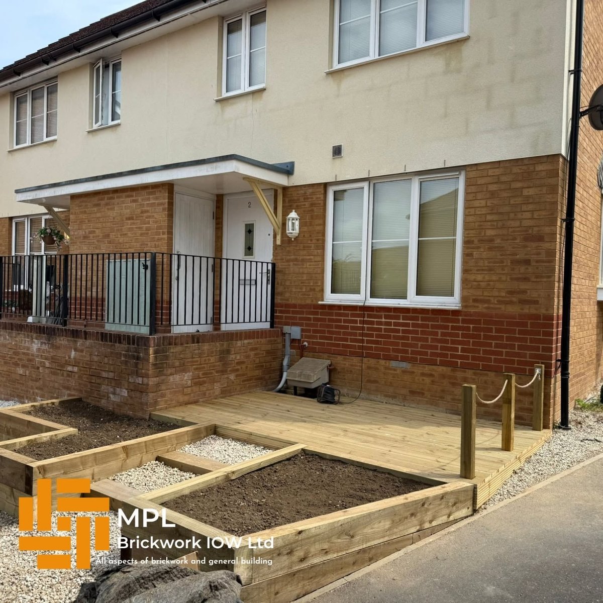 Modern two-story brick house extension with metal railings, raised wooden deck, and landscaped garden beds