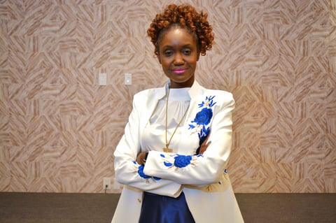 Woman with curly reddish-brown hair wearing a white blazer with blue floral design over a blue dress, smiling with arms crossed against a patterned beige wall