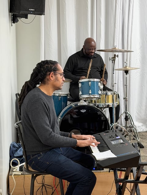 Two musicians in a studio: a person seated at a keyboard and another standing behind a blue drum kit during a recording session