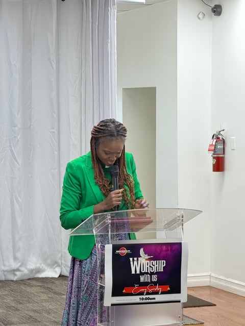 Woman in green blazer speaking at podium with Worship with Us sign in modern church sanctuary