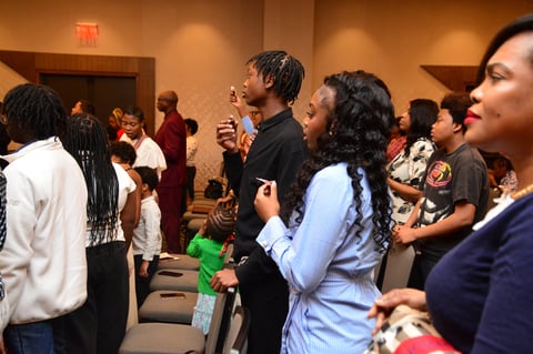 A diverse group of people gathered in an indoor venue with warm lighting, many standing and socializing during what appears to be a community or religious event