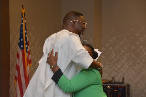 Man in white coat and glasses hugging woman in green shirt, with American flag visible in background