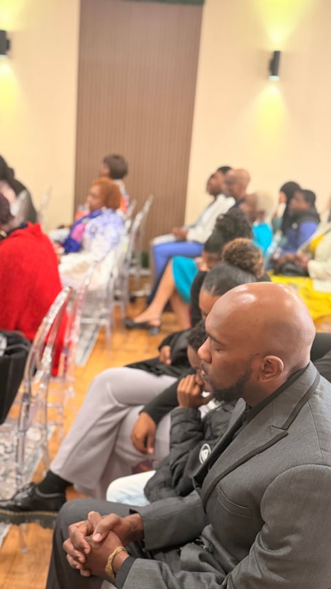 Man in gray suit sitting attentively at an indoor conference or community event with multiple attendees in the background