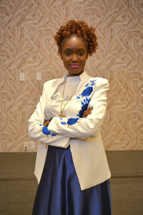 Woman with reddish-brown curly hair wearing a cream blazer with blue floral embroidery over a navy skirt, standing with arms crossed against a beige patterned wall