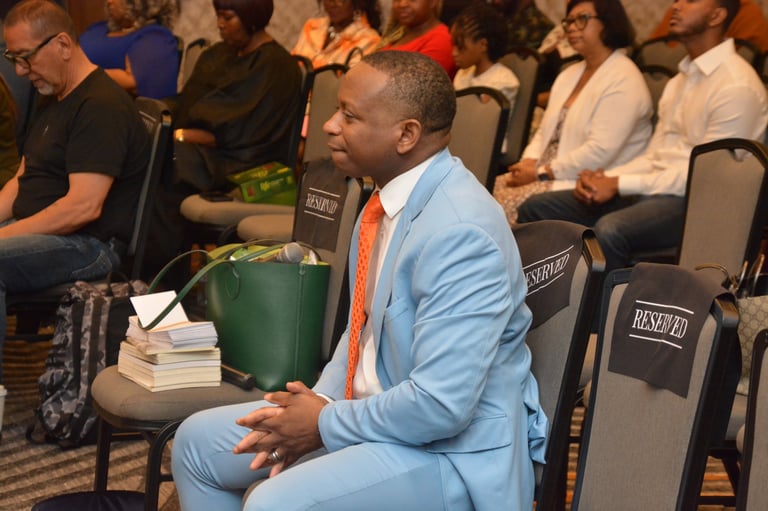 Man in light blue suit and orange tie sitting among audience members at an indoor event or conference