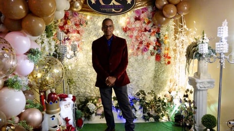Man in burgundy blazer standing in front of ornate floral backdrop with pink and white flowers, balloons, and decorative candelabras