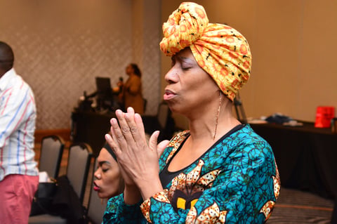 Woman wearing a gold turban and blue patterned shirt with hands clasped in prayer at an indoor gathering