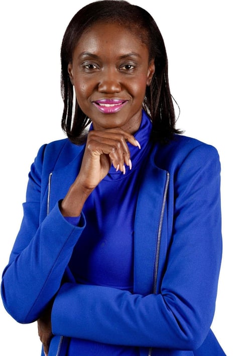 Professional portrait of a woman with long dark hair wearing a blue jacket, smiling with her hand near her chin against a white background