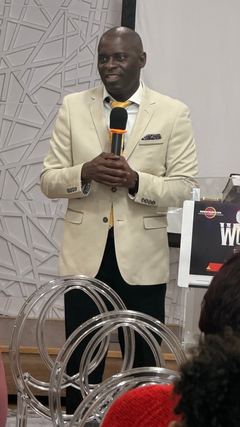 Man in cream blazer holding microphone while speaking at an event in front of an audience