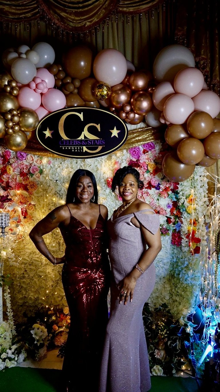Two women posing at a luxury event photo backdrop decorated with colorful flowers, gold balloons, and a Cs Stars & Stripes logo sign