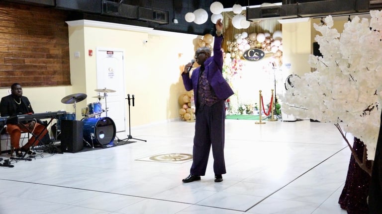 Person in dark purple outfit performing with arms raised in a studio space with band setup and decorations in background