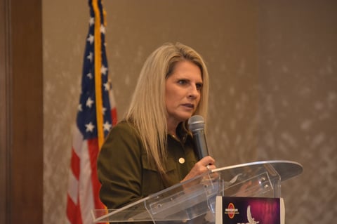 Woman with blonde hair speaking at a podium with microphone in front of an American flag