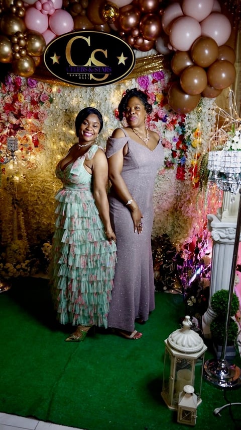 Two women posing at an elegant photo booth with floral backdrop, gold accents, and decorative balloons