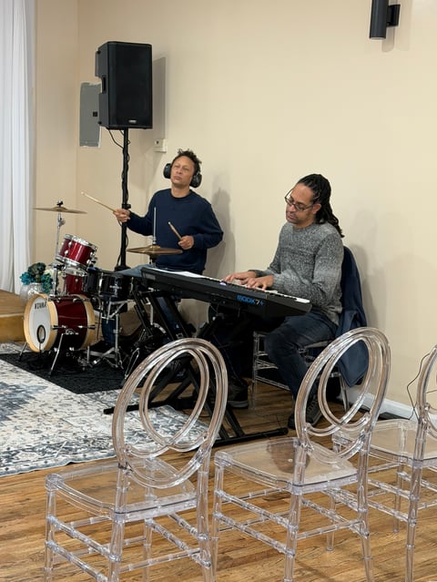 Two young musicians performing together - a child playing drums while a teenager plays keyboard in an indoor venue with empty chairs in foreground