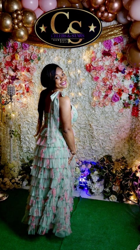 Woman in flowing multicolored tiered gown posing at elegant floral backdrop with gold balloons and ornate signage
