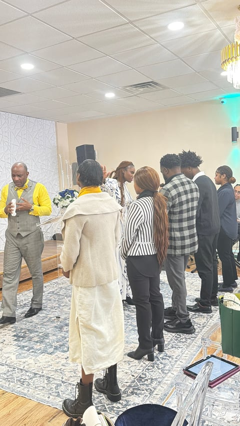 A group of people gathered in an indoor venue, listening to a man in a yellow jacket speaking during what appears to be a formal event or networking gathering