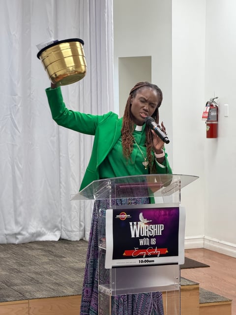 Woman in green jacket holding a gold bowl aloft while speaking at a clear podium labeled Worship with us