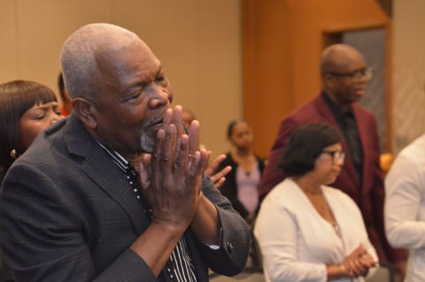 Older man with hands clasped near face in contemplative pose during indoor gathering