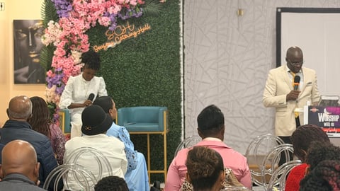 Man in beige suit speaking at podium to seated audience in front of pink floral backdrop and green moss wall