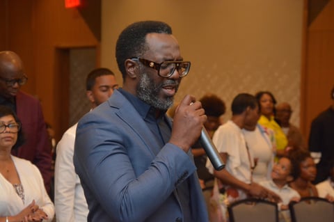 Man in blue blazer and glasses speaking into microphone at a community event with seated audience members in background