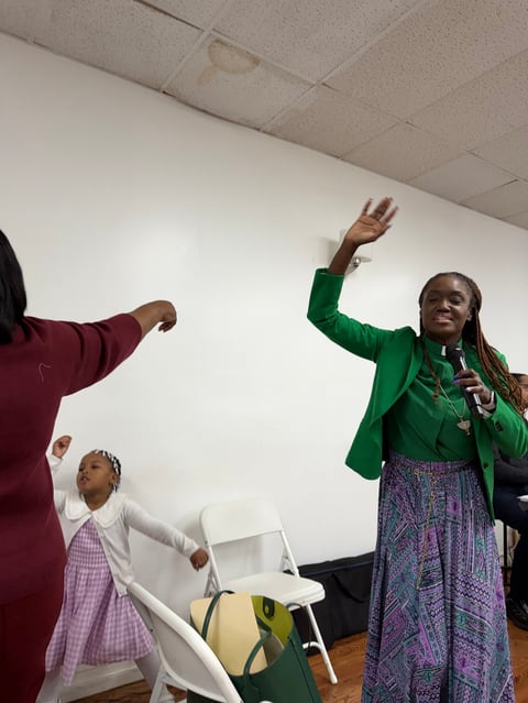 Woman in green jacket and purple skirt raises fist while speaking indoors with others present