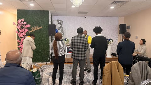 Group of people standing in an indoor venue with green wall and pink flowers, watching a presentation or event