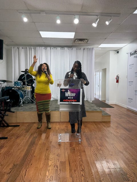 Two women performing on stage at a worship event, one in yellow shirt and one in patterned dress, with microphones and drum kit visible