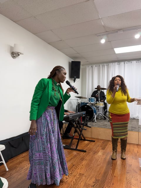 Two women performing on stage with microphones, one in green jacket and patterned skirt, the other in yellow top with colorful striped skirt, with band members in background